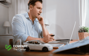 A man looks thoughtfully at a laptop screen, with a white model car on the desk in the foreground, representing the process of researching or buying a car online. The Compare The Carrier logo is visible.